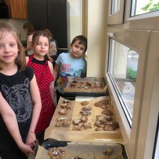 Na zdjęciu widzę grupę dzieci w pomieszczeniu o dużych oknach, przez które wpada naturalne światło. Niektóre z dzieci mają na sobie fartuchy, co sugeruje że odbywają się warsztaty kulinarne. Na parapecie leżą na blasze ciasteczka.