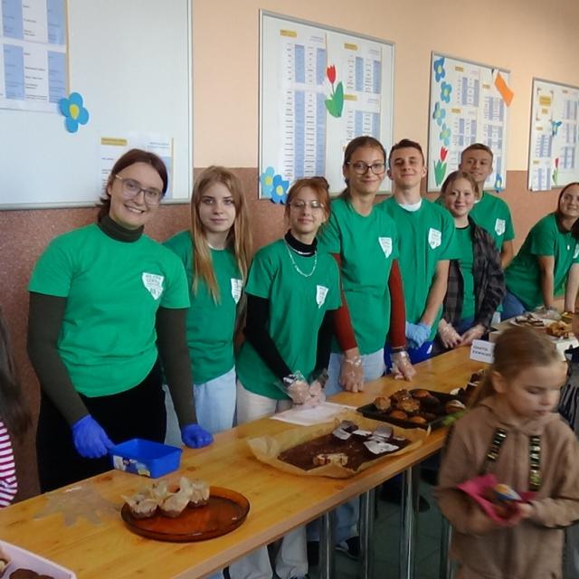 Grupa wolontariuszy z MSP2 w zielonych koszulkach za stołami z wypiekami.