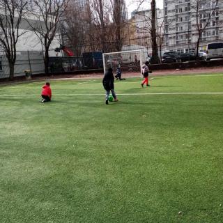 Uczniowie zerówki grają w piłkę na boisku szkolnym. Biegają po zielonej murawie, w tle widać bramkę i blok mieszkalny za ogrodzeniem szkoły.