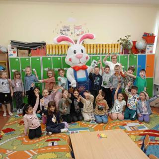 Uczniowie w klasie pozują do zdjęcia z osobą przebraną w strój wielkiego pluszowego królika. Niektóre dzieci siedzą na kolorowym dywanie, inne stoją. W tle widać szafki uczniów w kolorze zielonym, pomarańczowym i niebieskim.