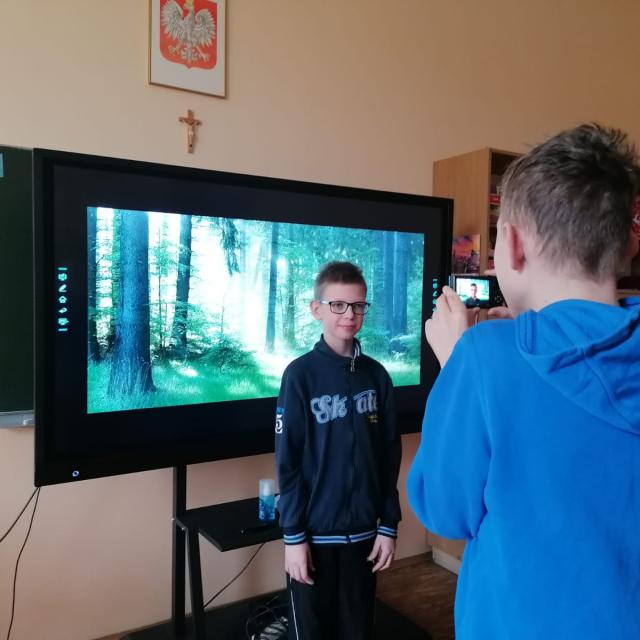 Chłopiec stojący tyłem, w bluzie z kapturem, robi zdjęcie koledze stojącemu na tle monitora interaktywnego. Nad monitorem wisi godło i krzyż.