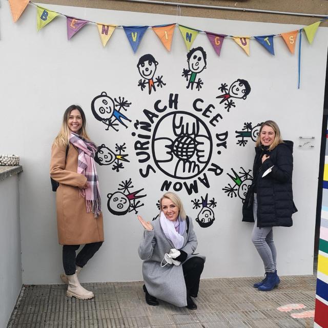 Trzy kobiety na tle logo hiszpańskiej szkoły partnerskiej. Dwie stoją, jedna kuca. Uśmiechają się.