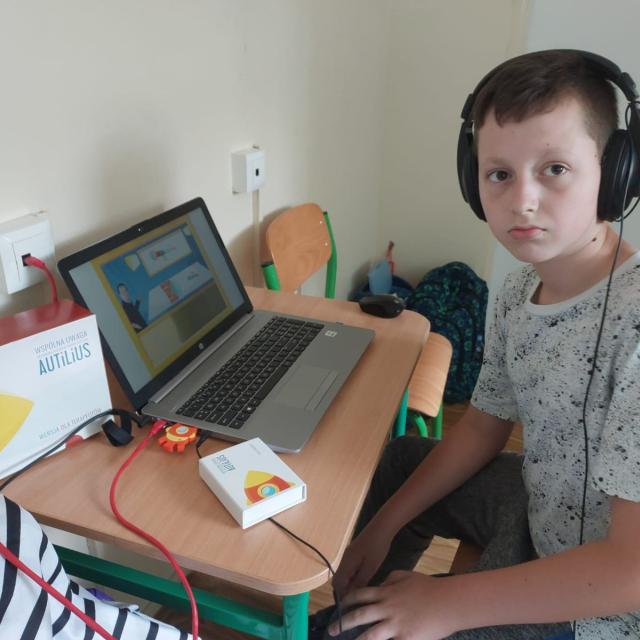 Uczeń ze słuchawkami na uszach siedzi przy stoliku, na którym jest laptop.